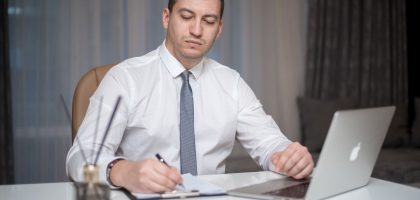 Professional businessperson in corporate attire writing at a white office desk with a laptop.