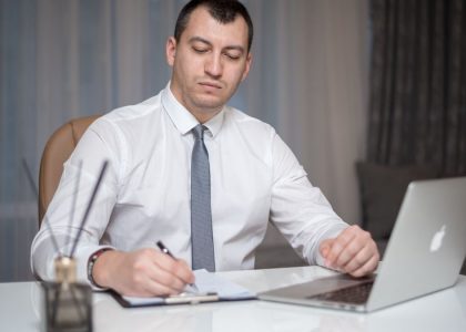 Professional businessperson in corporate attire writing at a white office desk with a laptop.